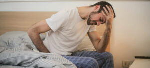 Exhausted man sitting at the edge of the bed, wearing pajamas, holding his left hand in his head, looking tired.