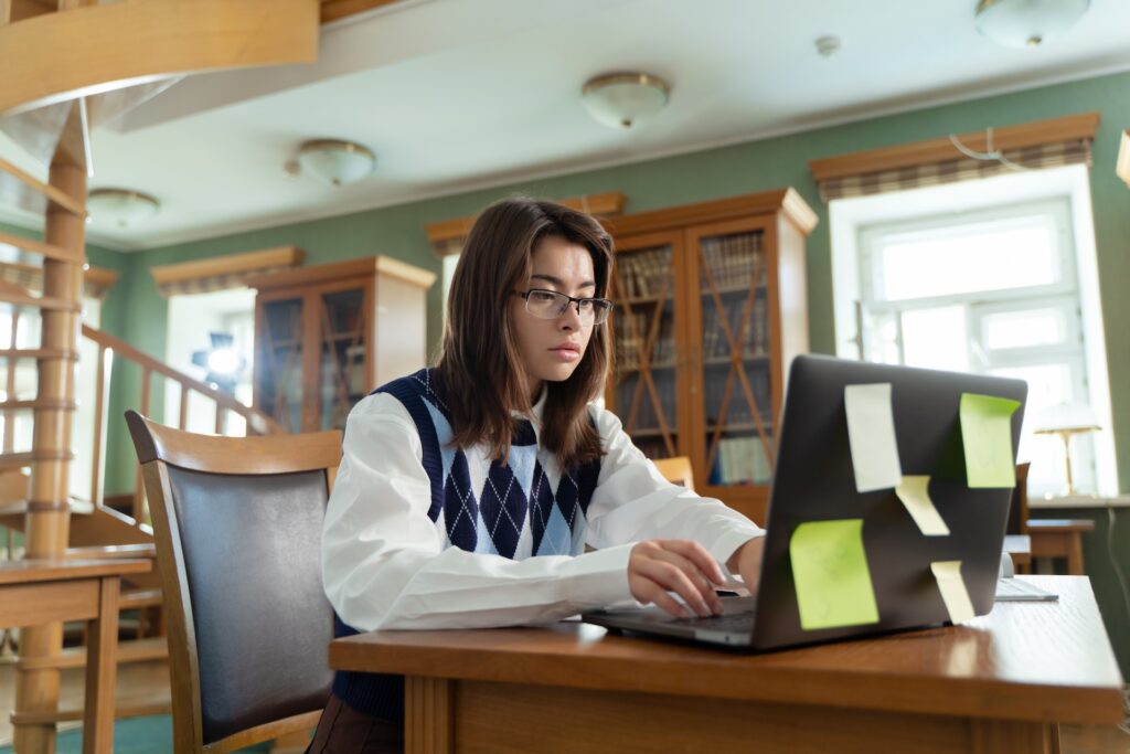 Female student studying at desk with her laptop, which has post-it notes. Inside a university library.